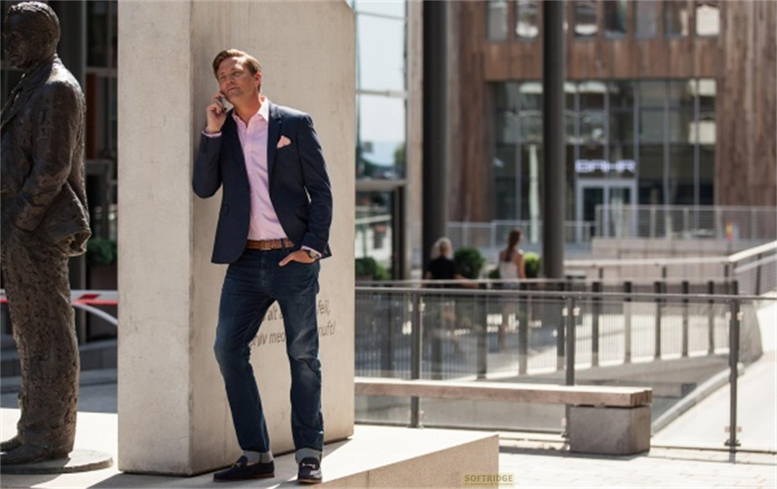 Uomo in giacca e jeans parla al telefono vicino a statua ed edificio.
