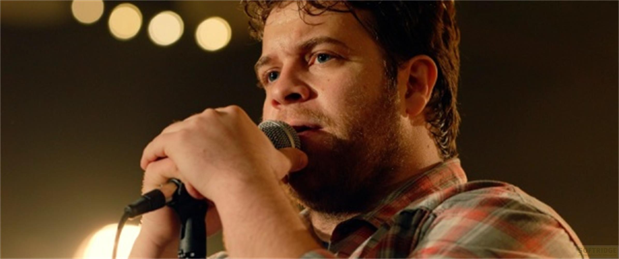 Un uomo con la barba canta in un microfono, luci sullo sfondo.