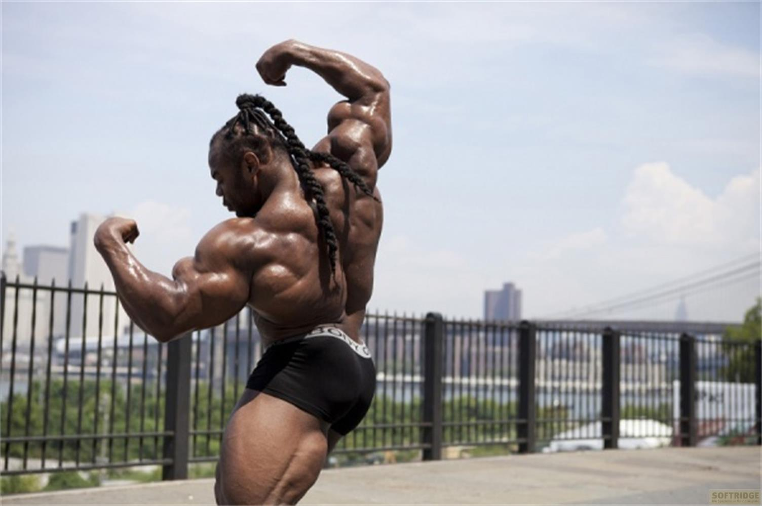Ein bodybuilder mit geflochtenem Haar posiert mit angespannten Muskeln vor dem Hintergrund einer Stadt.