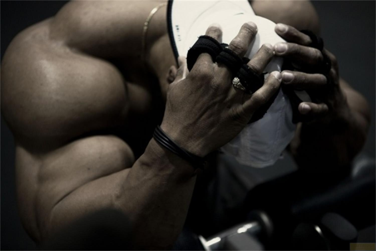 Muskulöser Mann im Fitnessstudio, trägt Handgelenkbandagen, hält eine weiße Kappe. Hanteln sichtbar.