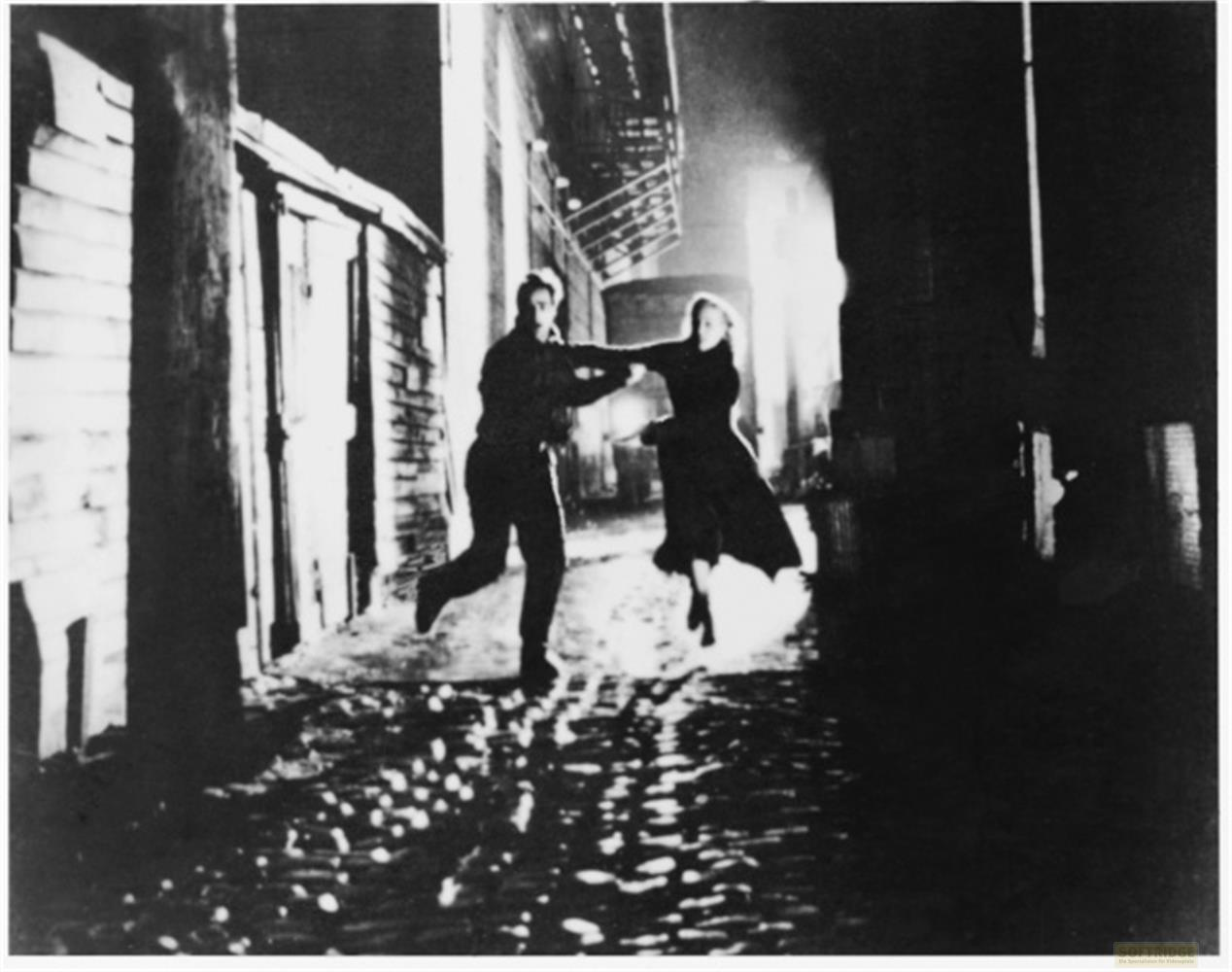Un homme et une femme dansent dans une rue sombre et mouillée entre des bâtiments.