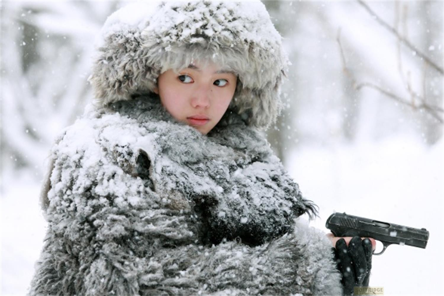 Una giovane donna in un folto cappotto di pelliccia con cappuccio tiene una pistola nella neve.