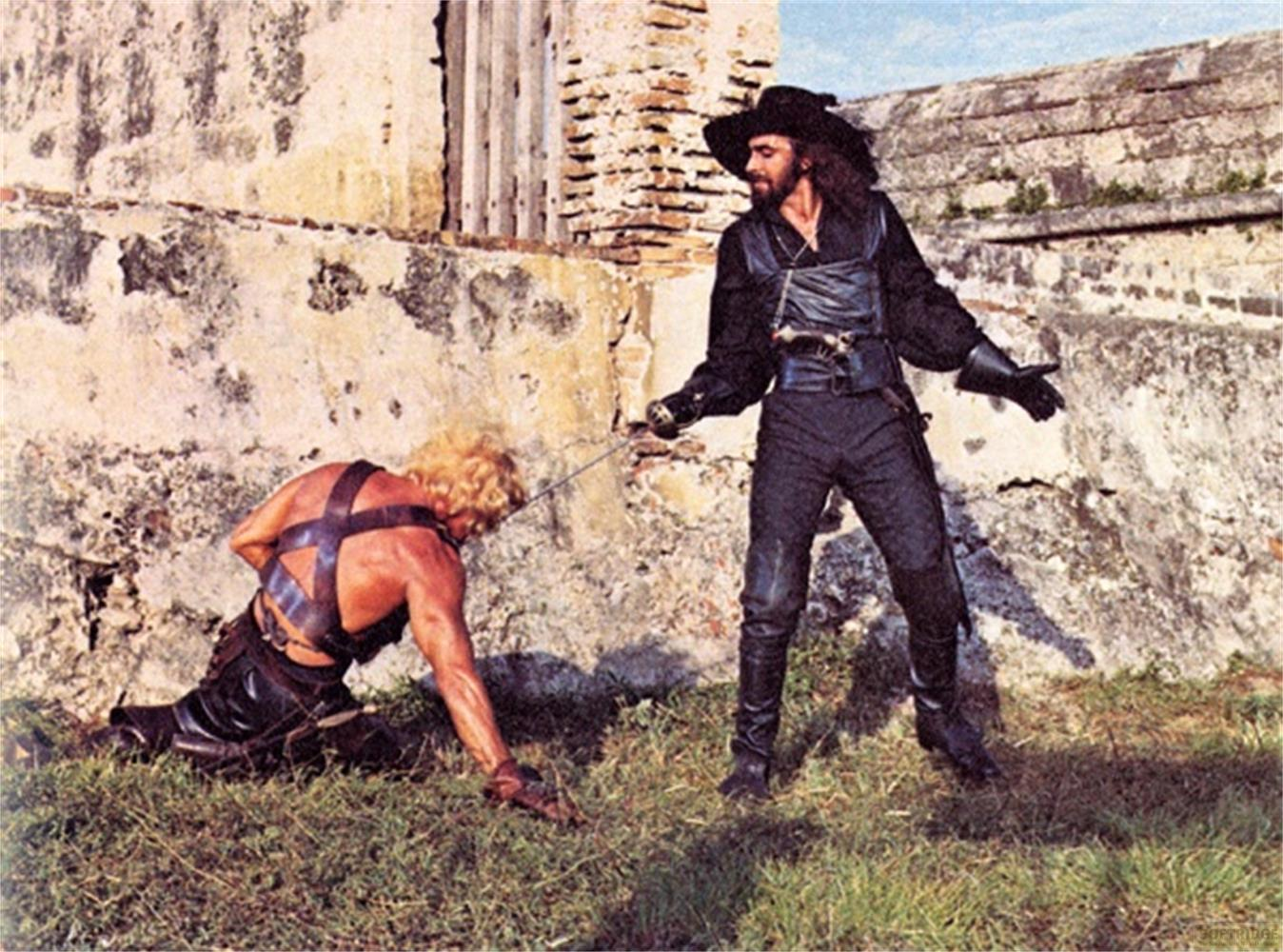 Un uomo con una spada minaccia un uomo a terra davanti a un muro.