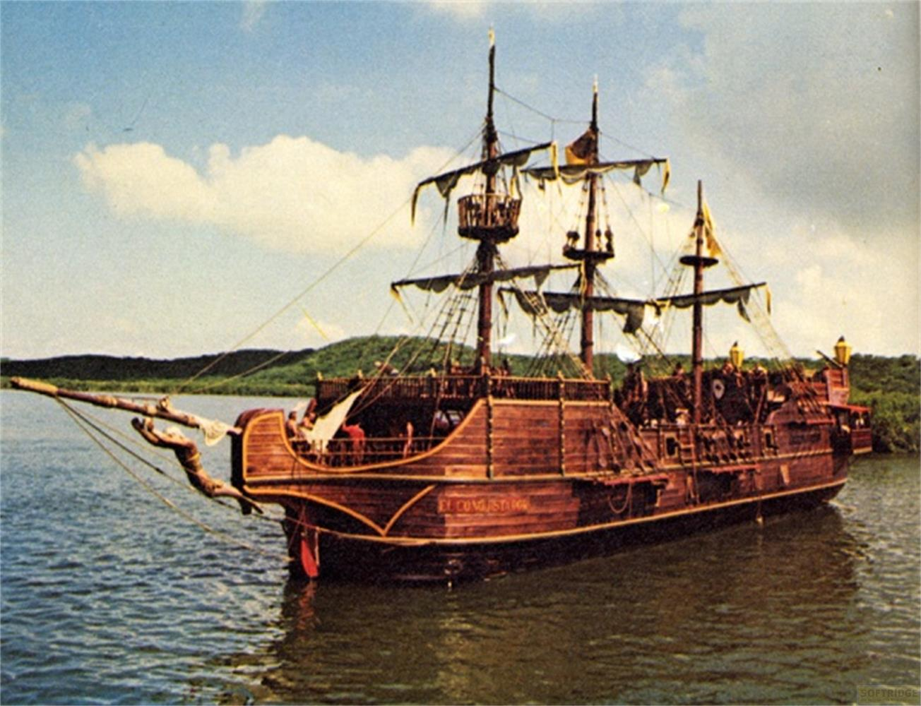 Una nave a vela in legno con più alberi galleggia sull'acqua, con una costa verde sullo sfondo.