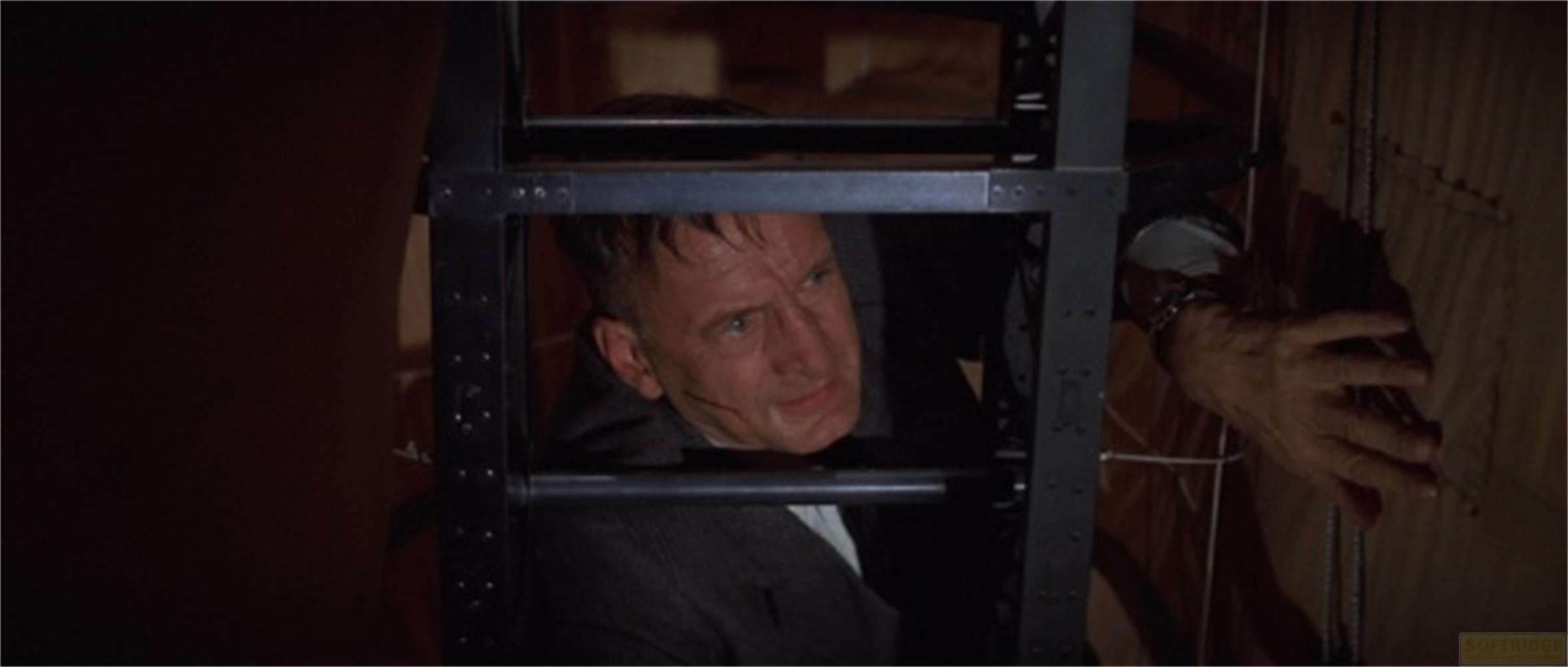 Homme en costume sombre grimpe dans une cage métallique, visage tendu.