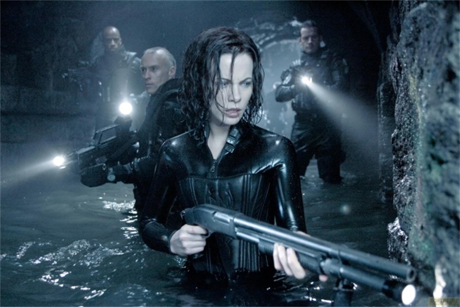 Une femme en tenue noire tient un fusil à pompe, debout dans l'eau. Des hommes armés de lampes de poche sont derrière.