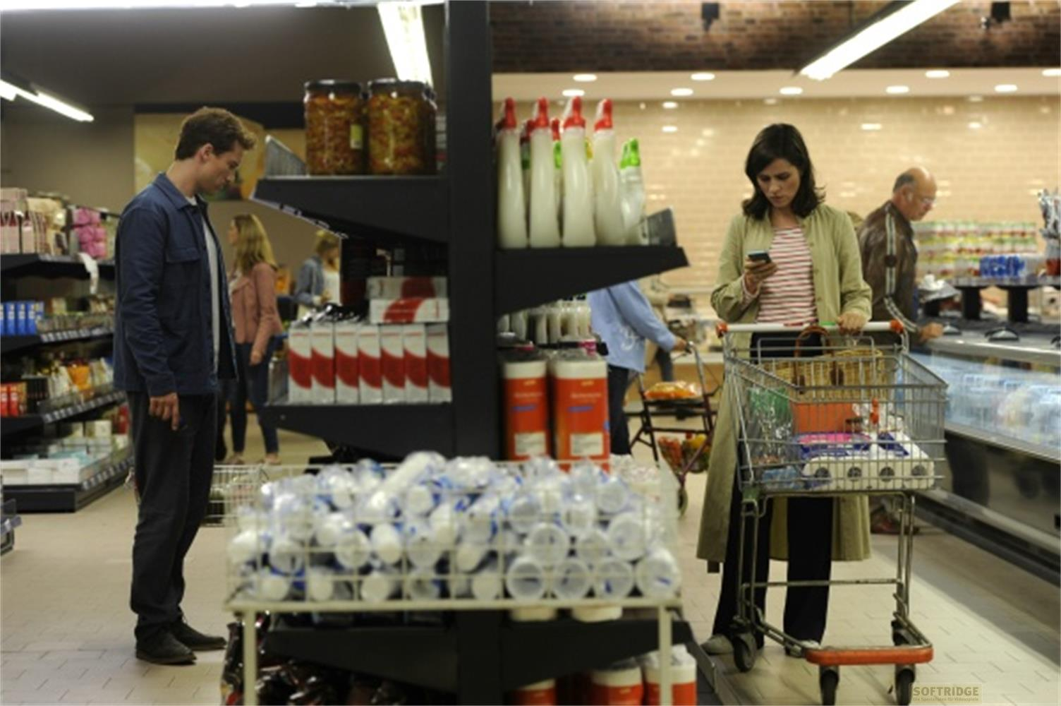 Persone che fanno la spesa in un supermercato, una donna guarda il telefono.
