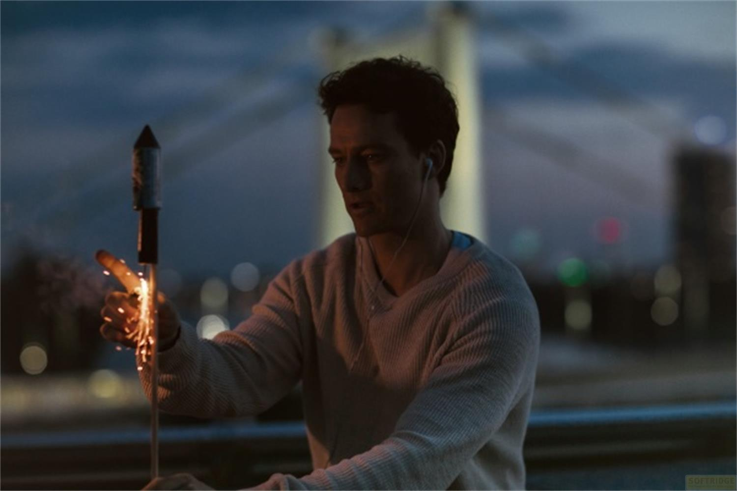 Uomo accende un petardo con scintille, indossa auricolari, luci della città sullo sfondo.