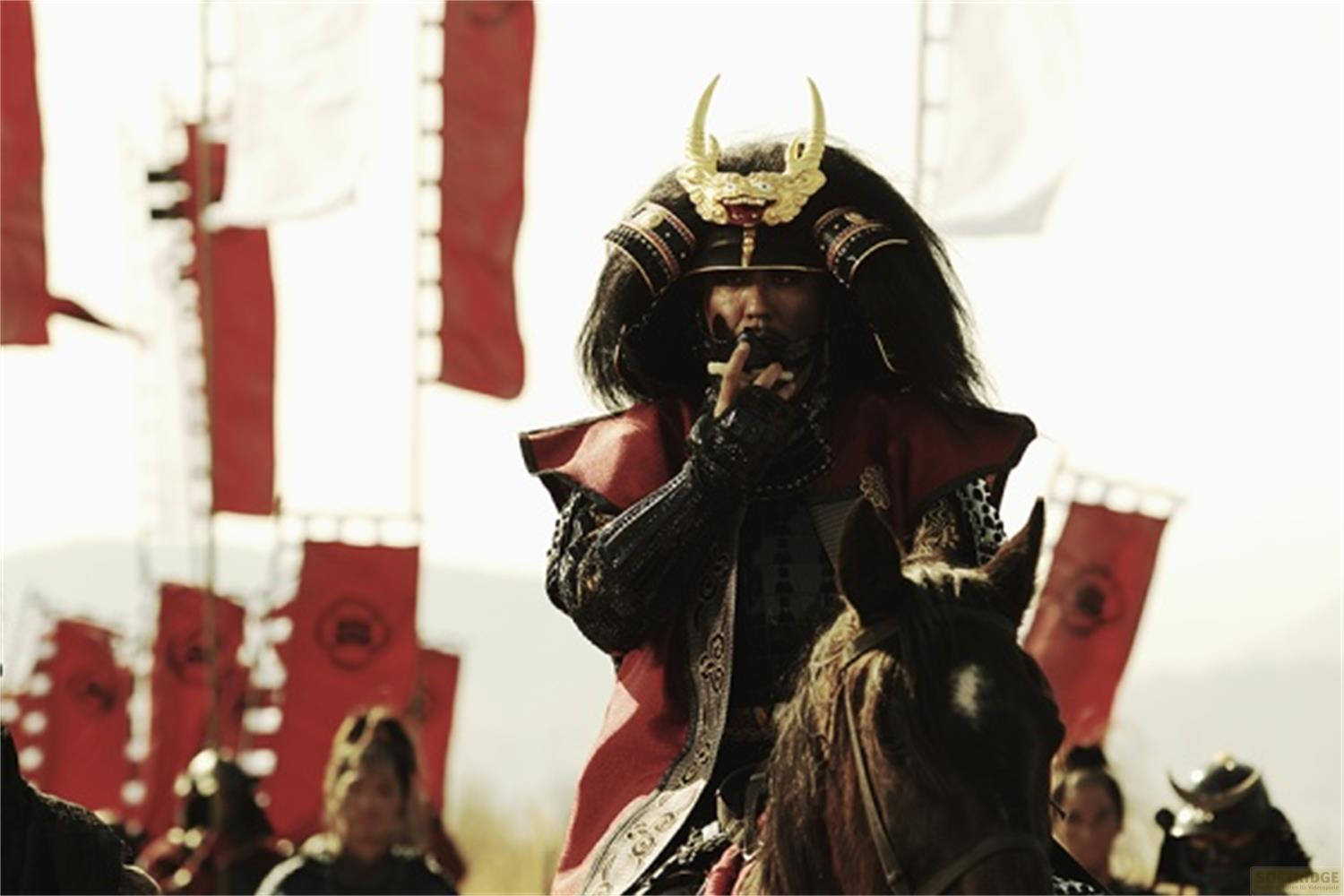 Un samouraï en armure complète monte à cheval, drapeaux rouges en arrière-plan.