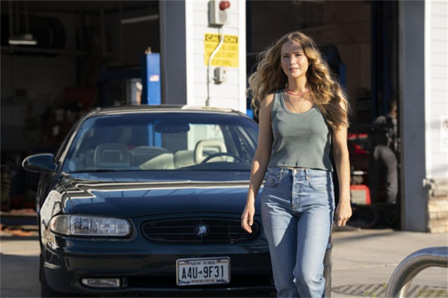 Donna in canottiera e jeans sta accanto a una berlina scura davanti a un garage.