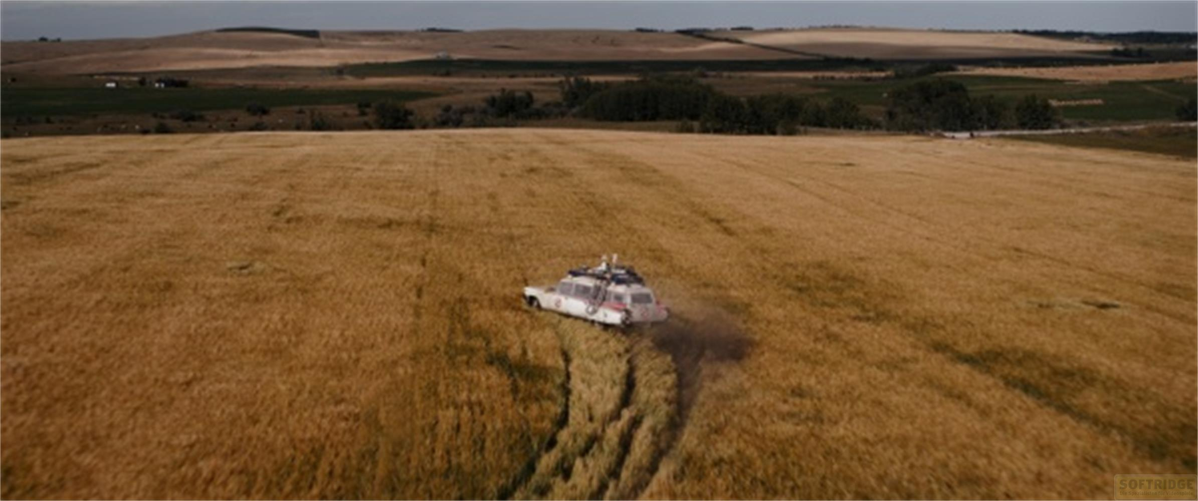 Ein Auto fährt durch ein Feld mit goldenem Getreide, Staub wirbelt auf.