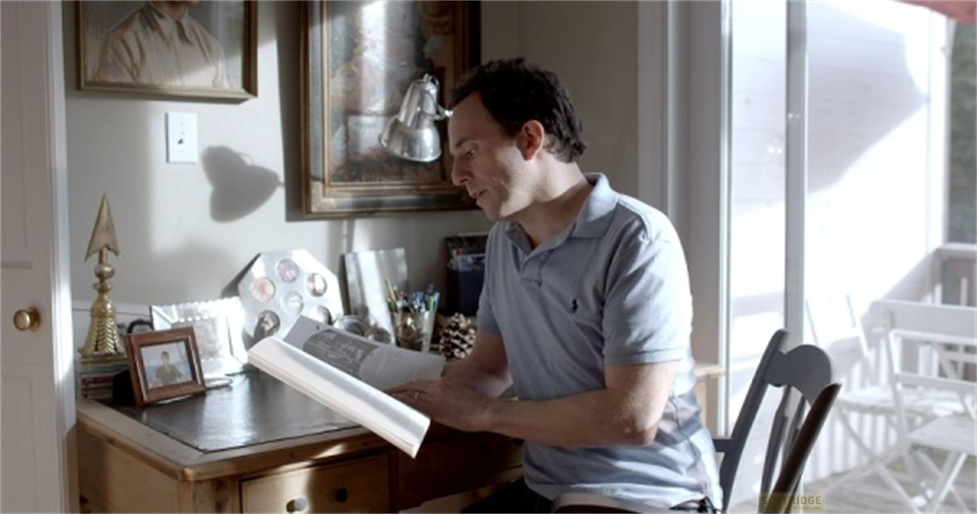 Un uomo siede a una scrivania, leggendo un libro. Scrivania con foto.