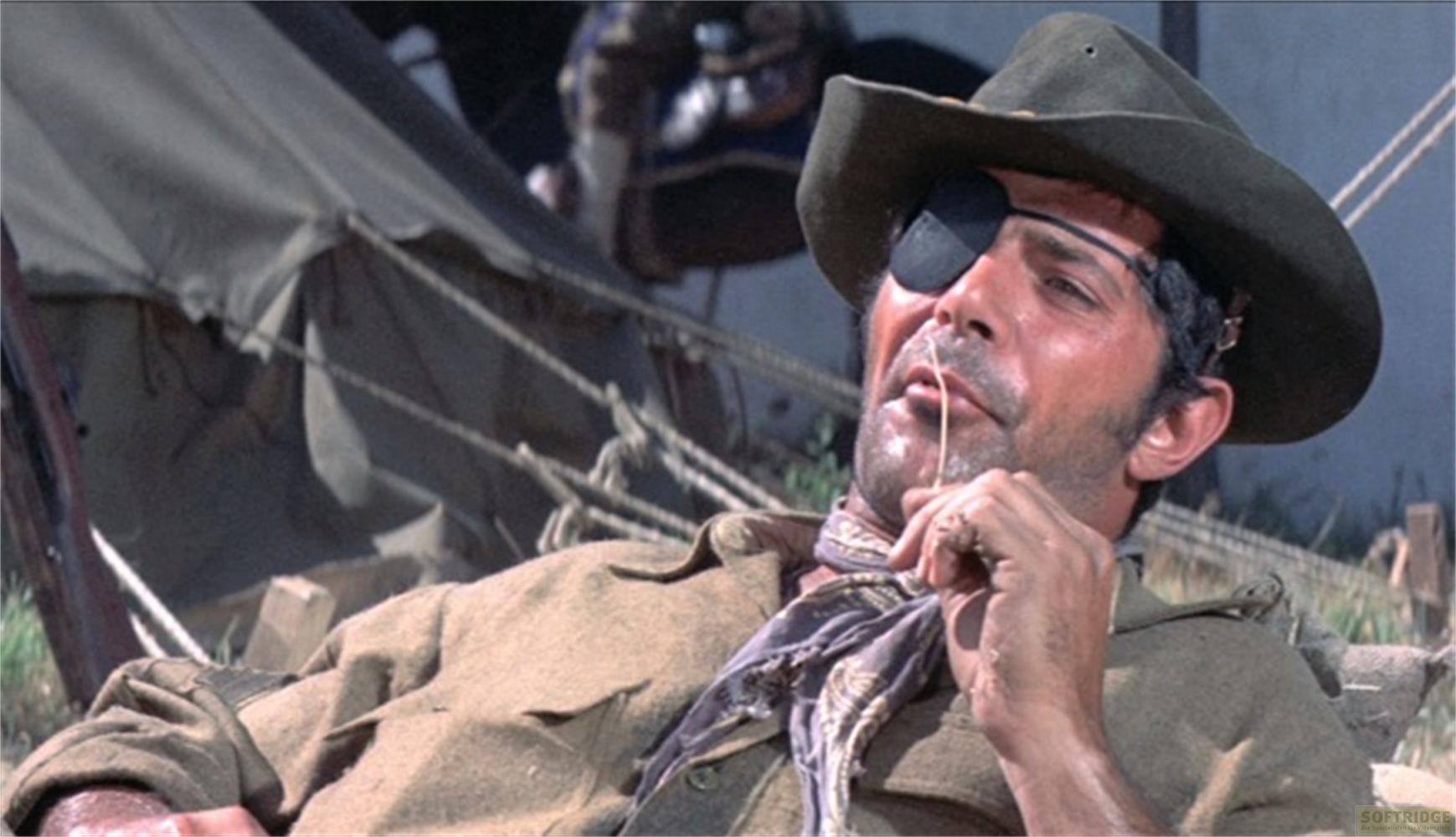 Un uomo con una benda sull'occhio indossa cappello e sciarpa. Sullo sfondo una tenda.