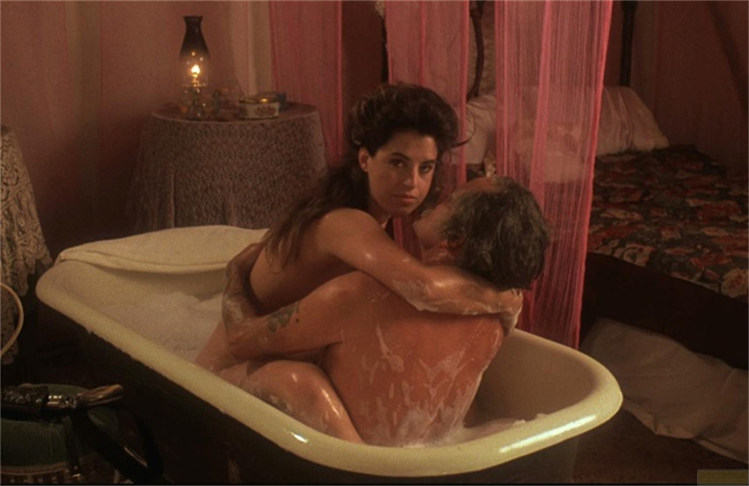 Donna e uomo in vasca da bagno schiumosa, letto sullo sfondo.