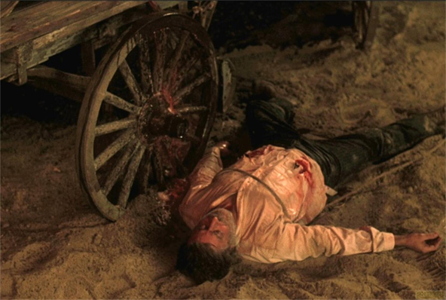 Un uomo giace sanguinante accanto alla ruota di un carro su terreno sabbioso.