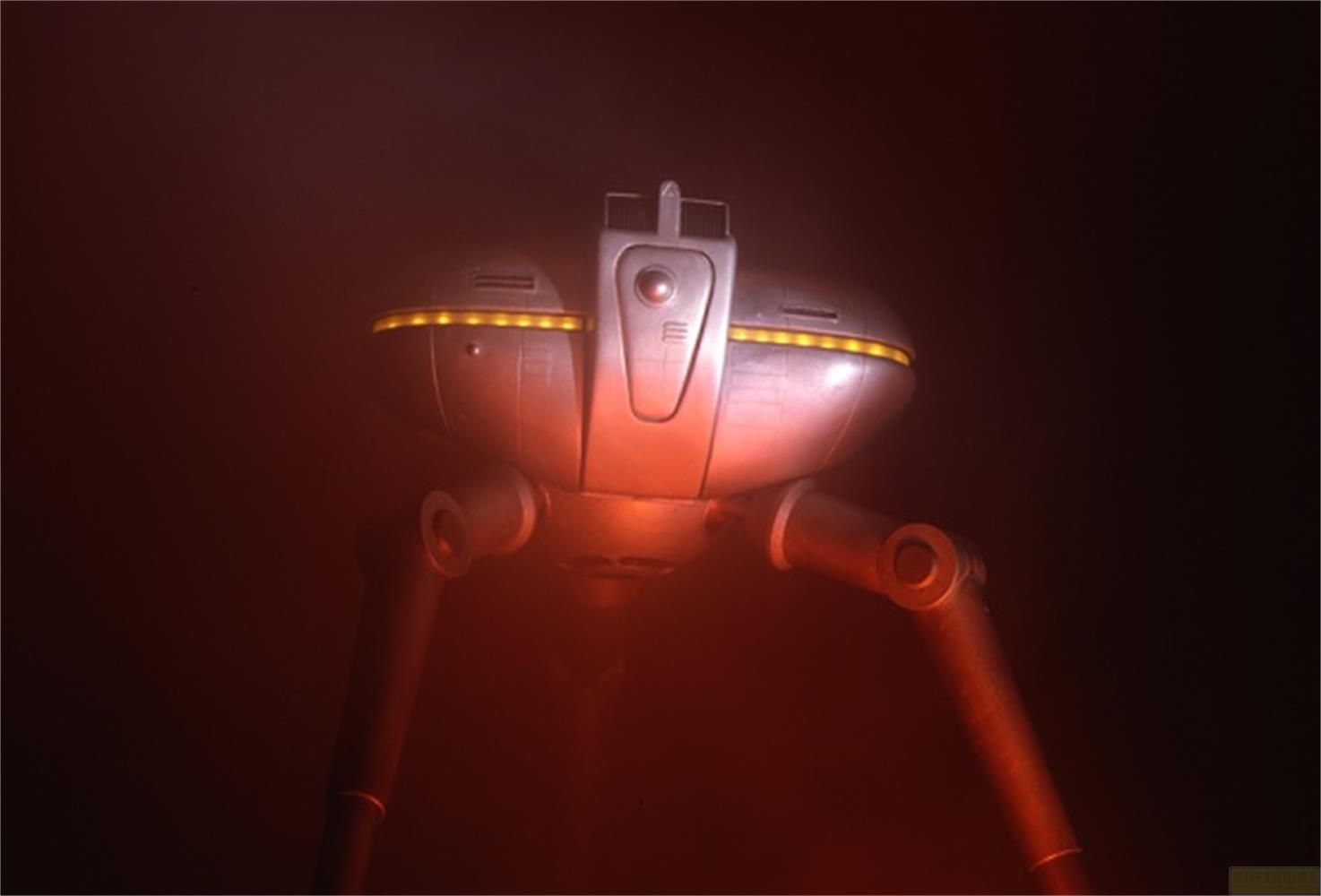 Robot a treppiede argentato con luci gialle, su sfondo scuro.