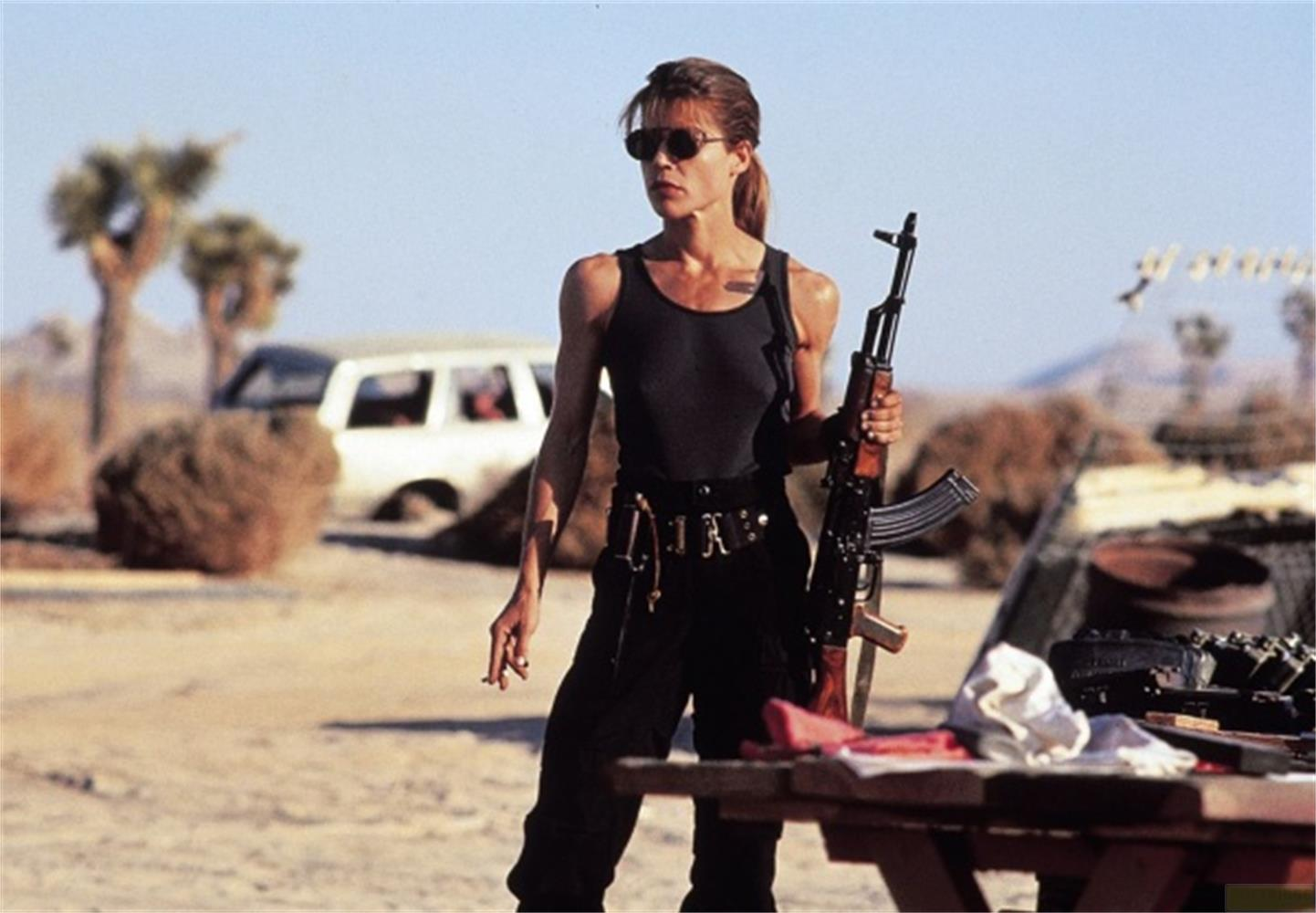 Eine Frau in Sonnenbrille und schwarzer Kleidung hält ein Gewehr in einer Wüstenlandschaft.
