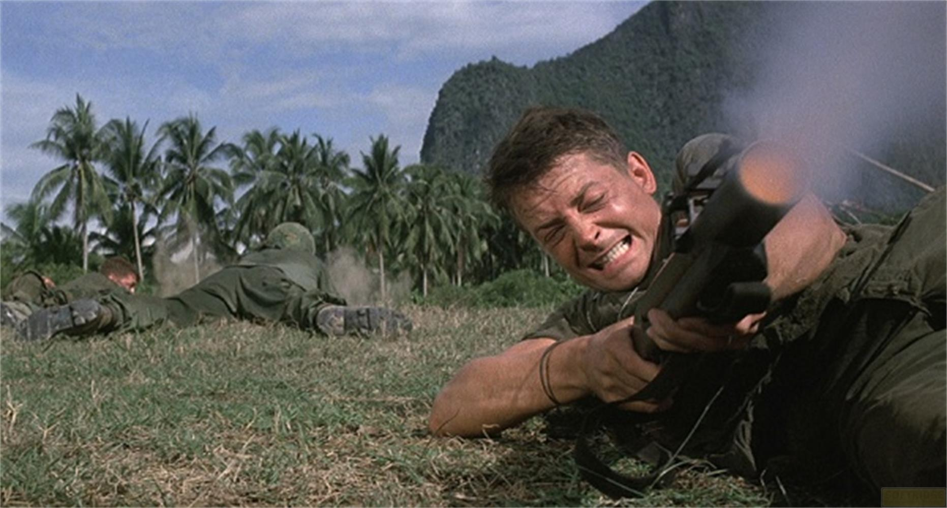 Soldats dans l'herbe, l'un tire une arme, palmiers et montagne en arrière-plan.