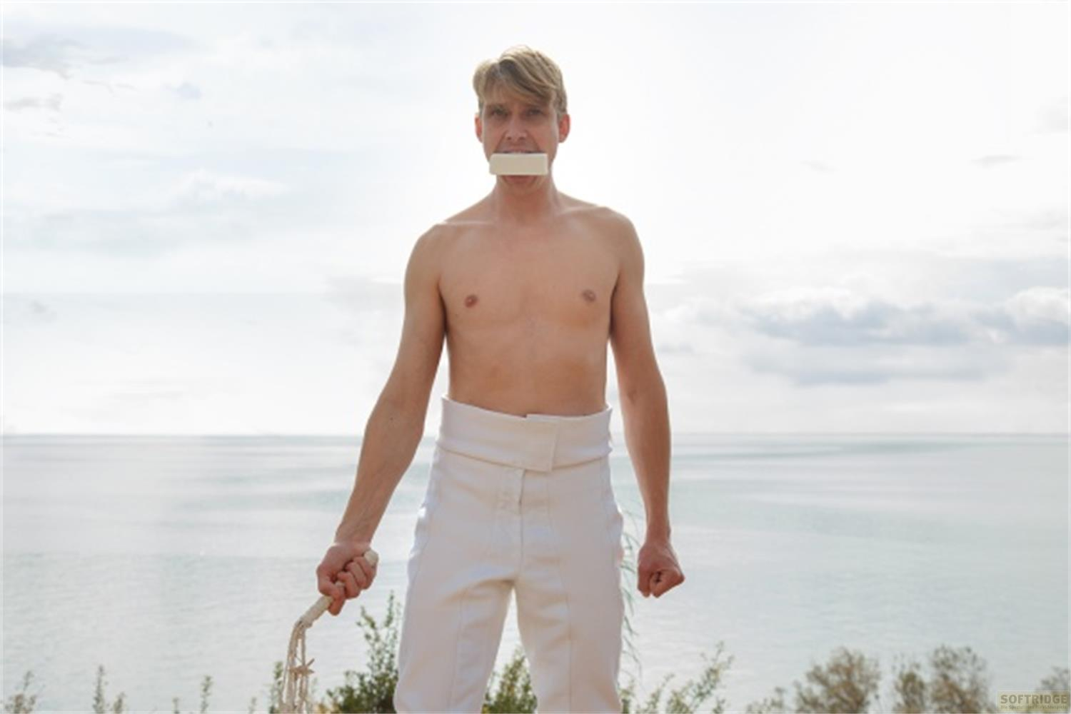Mann mit Band um den Mund und Seil in der Hand am Meer.