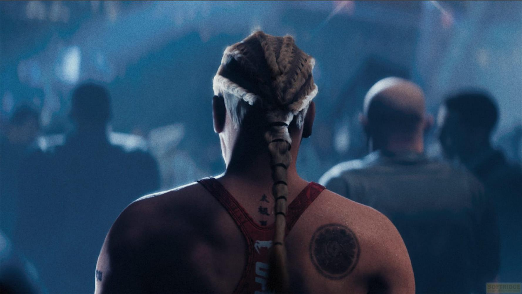 Widok z tyłu mężczyzny z zaplecionymi włosami w czerwonym podkoszulku. Niewyraźne sylwetki w tle.