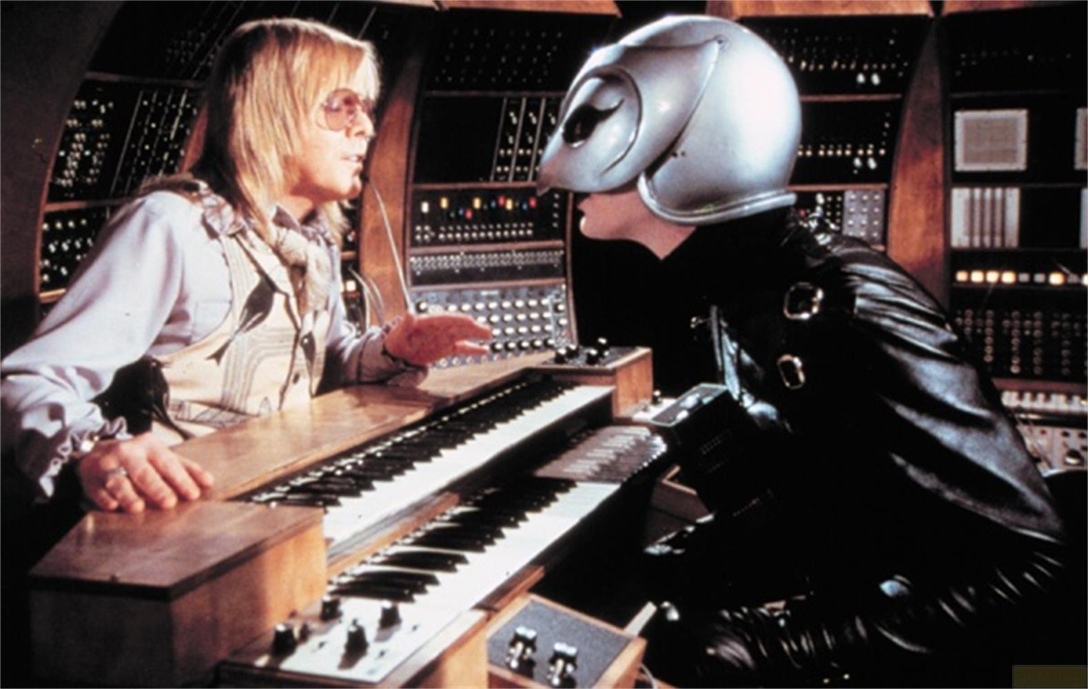 Ein Mann mit Helm spricht mit einem Mann mit langen Haaren an einem Keyboard.