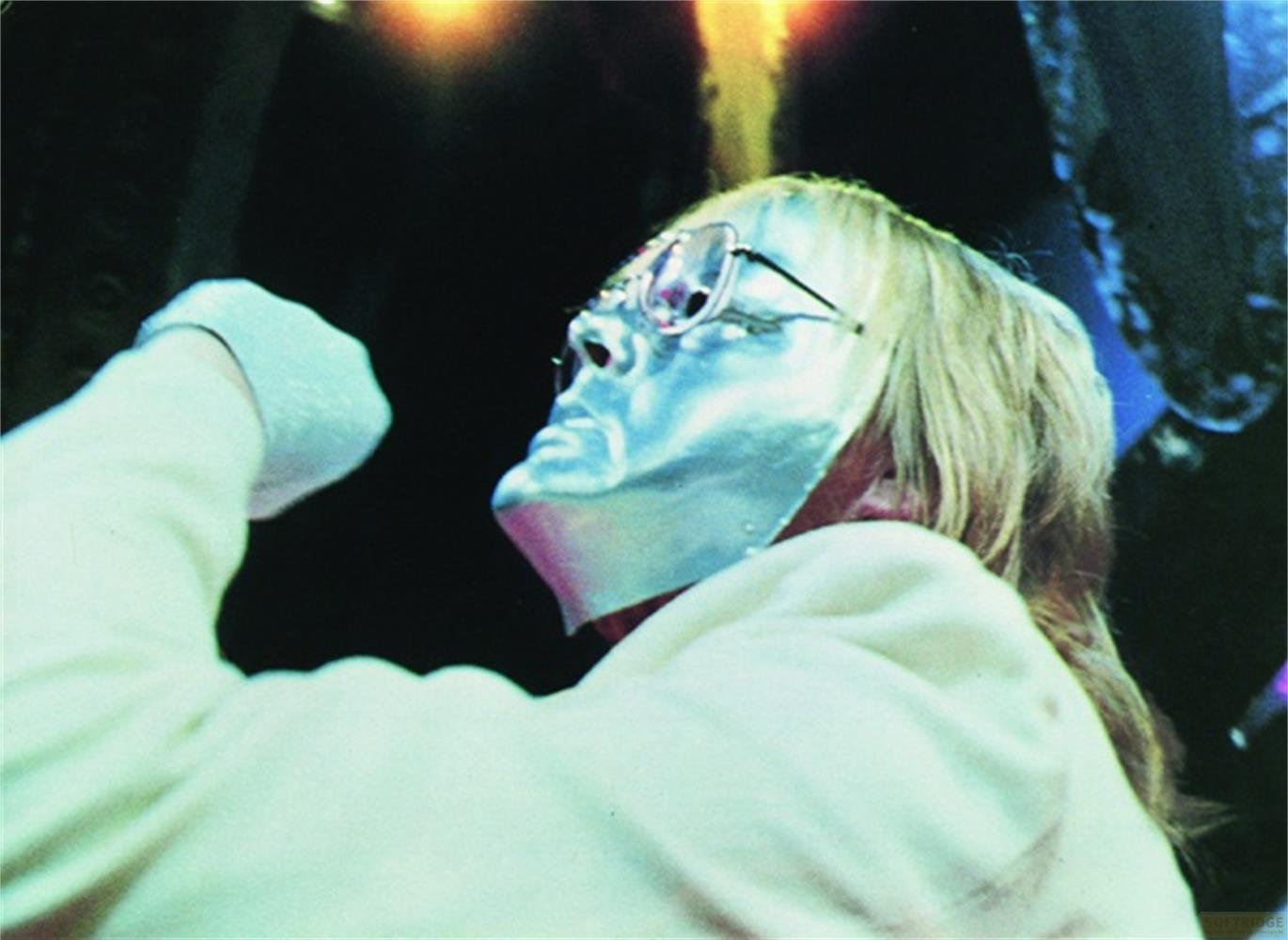 Person mit silberner Maske und Brille, weißer Pullover, im Dunkeln.