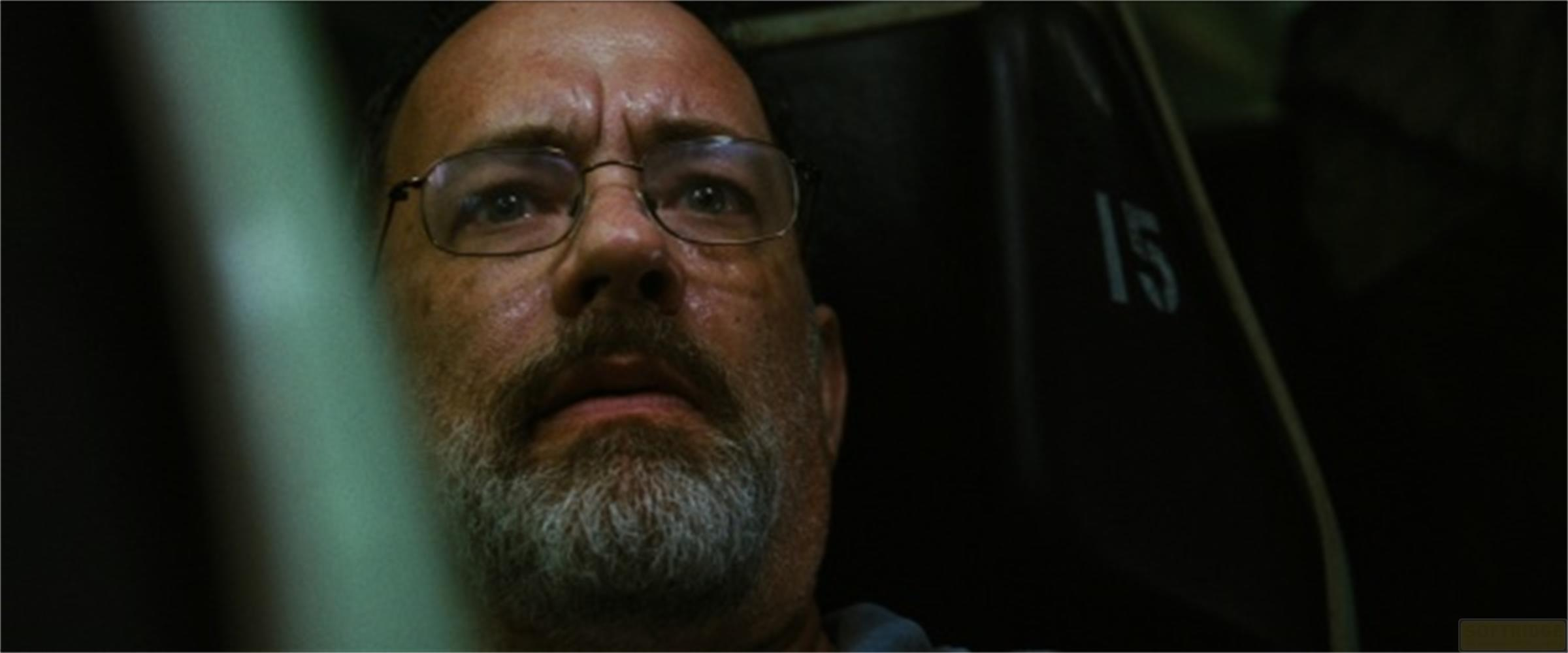 Un homme avec des lunettes et une barbe regarde la caméra avec inquiétude. Il est dans une pièce sombre.