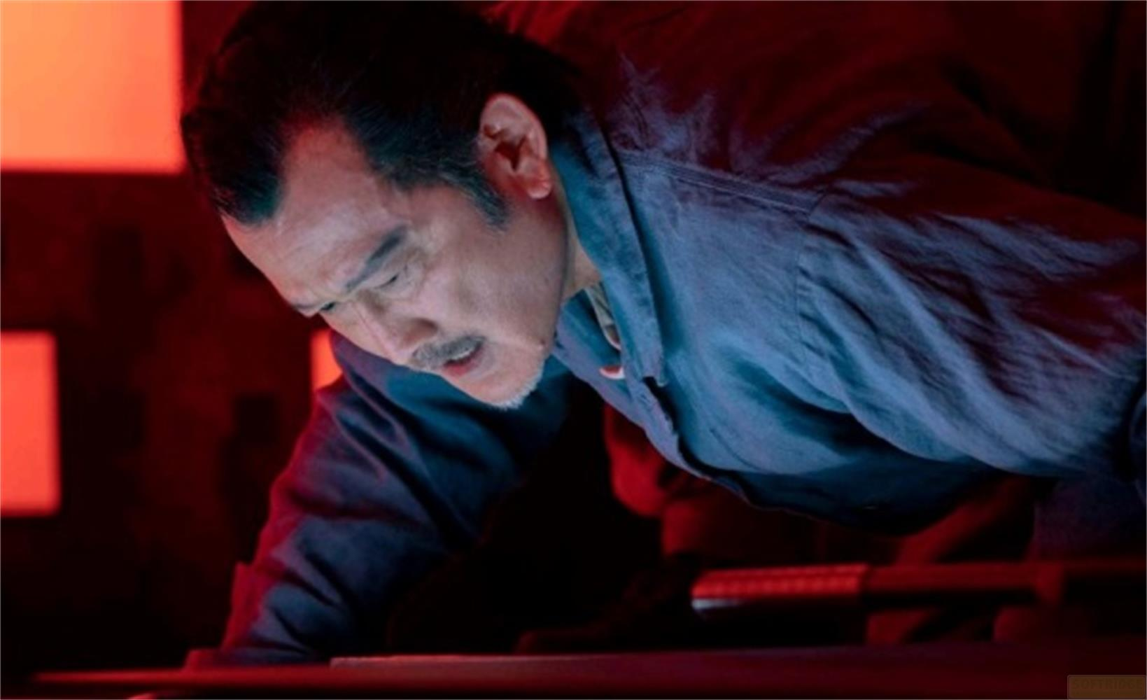 Un uomo con i baffi si china su un tavolo, con luce rossa sullo sfondo.