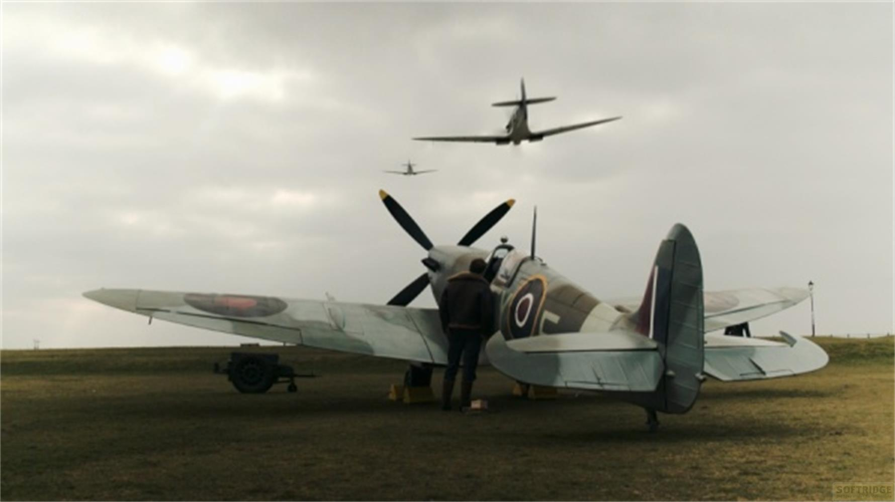 Due uomini accanto a un aereo Spitfire sull'erba. Un altro aereo vola sopra.