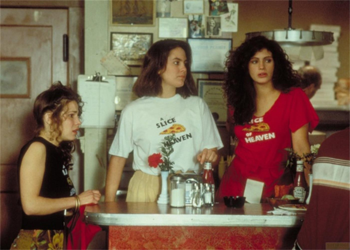 Tre donne al bancone. Due indossano magliette con grafiche di pizza.