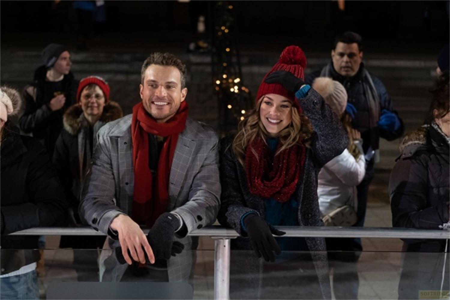 Un uomo e una donna sorridenti in abiti invernali in primo piano, luci colorate sullo sfondo.