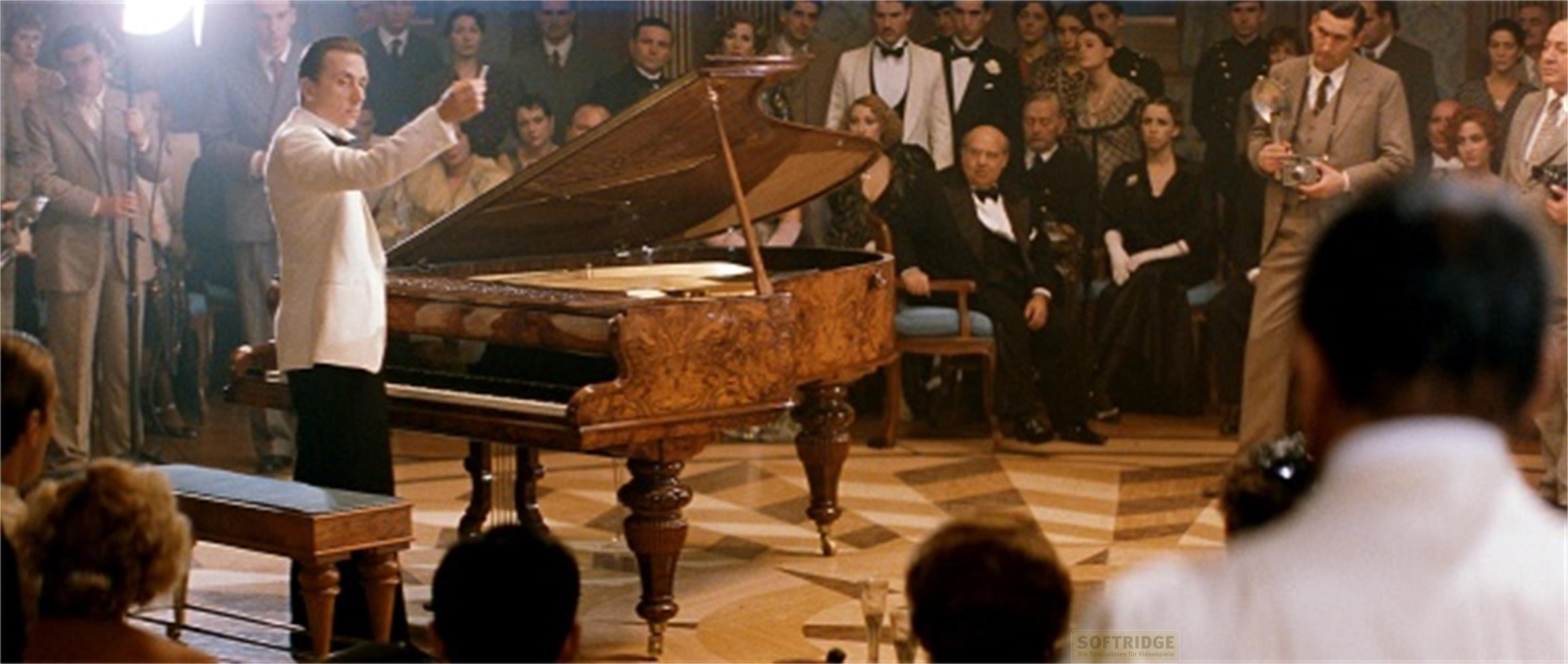 Un uomo in giacca bianca dirige davanti a un pianoforte a coda. Spettatori in piedi intorno.