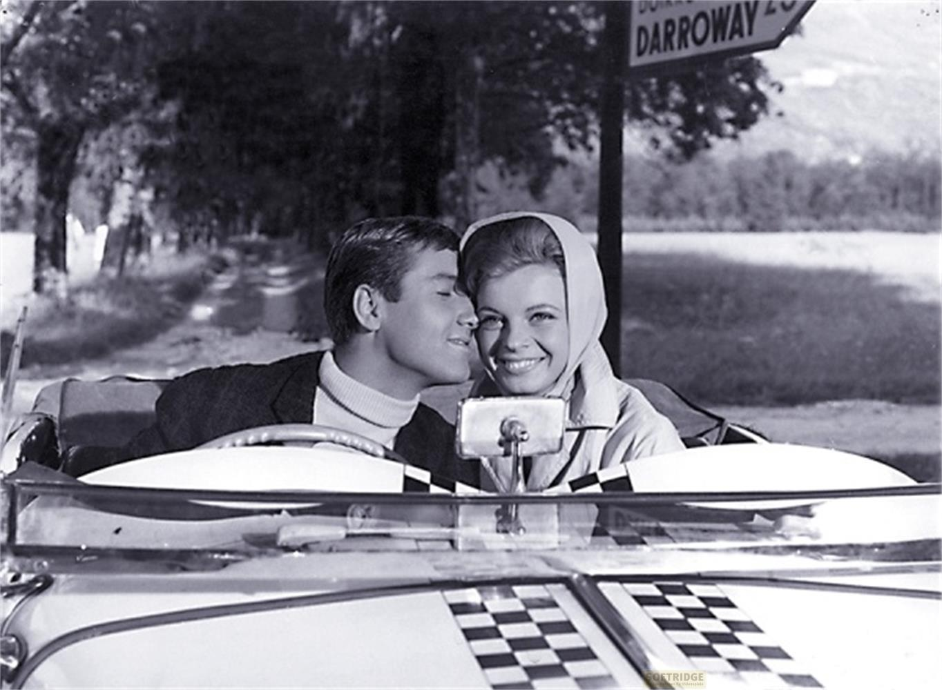 Un homme murmure à l'oreille d'une femme souriante dans une voiture décapotable.