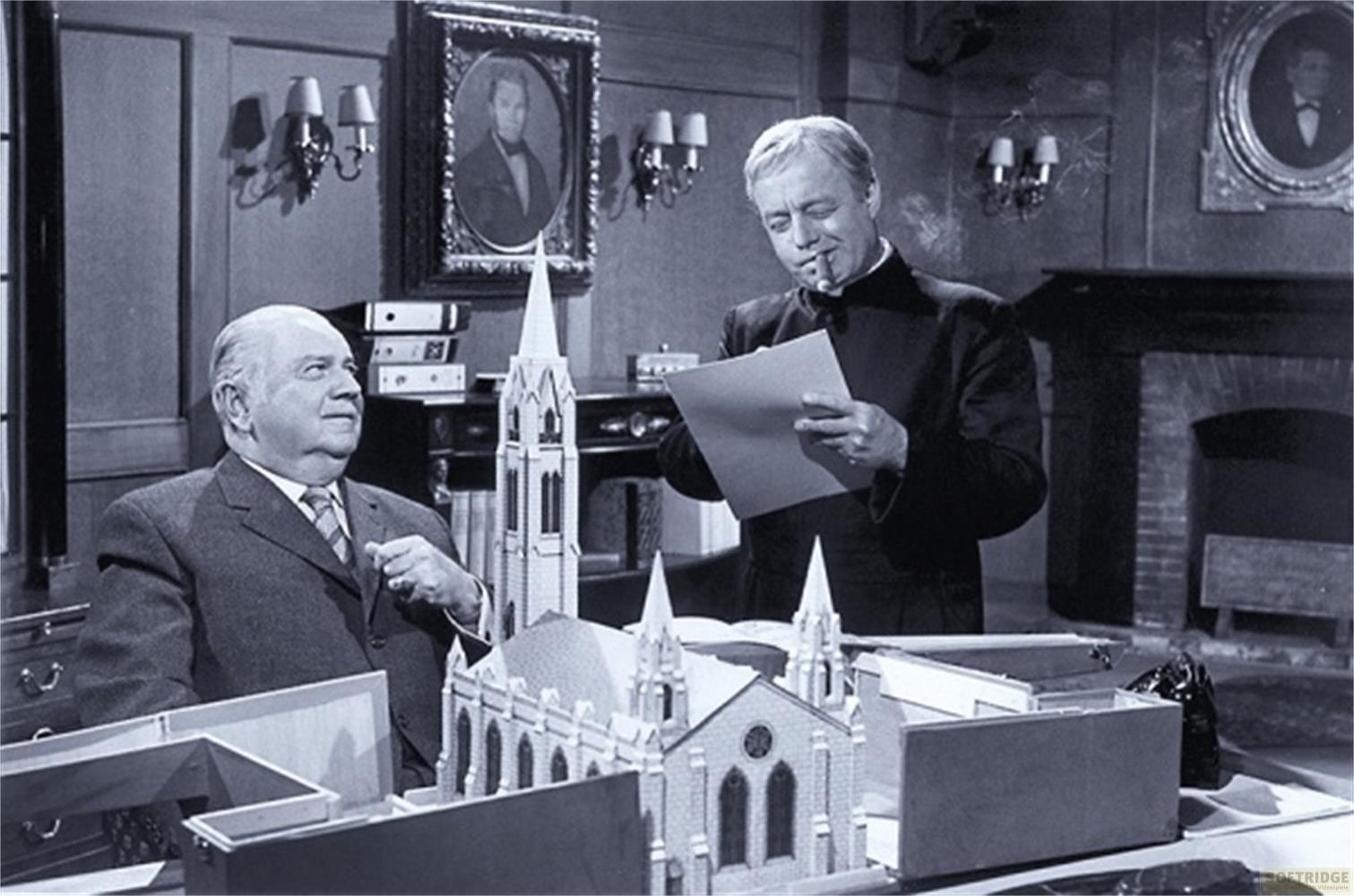 Deux hommes dans une pièce avec des maquettes d'églises et des portraits au mur.