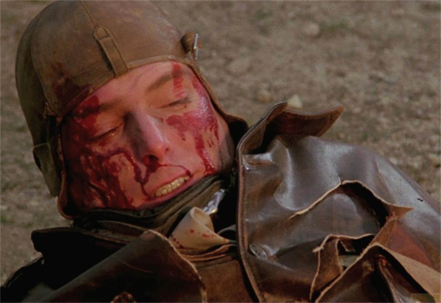 Un pilota con il viso insanguinato e il casco di pelle giace a terra.