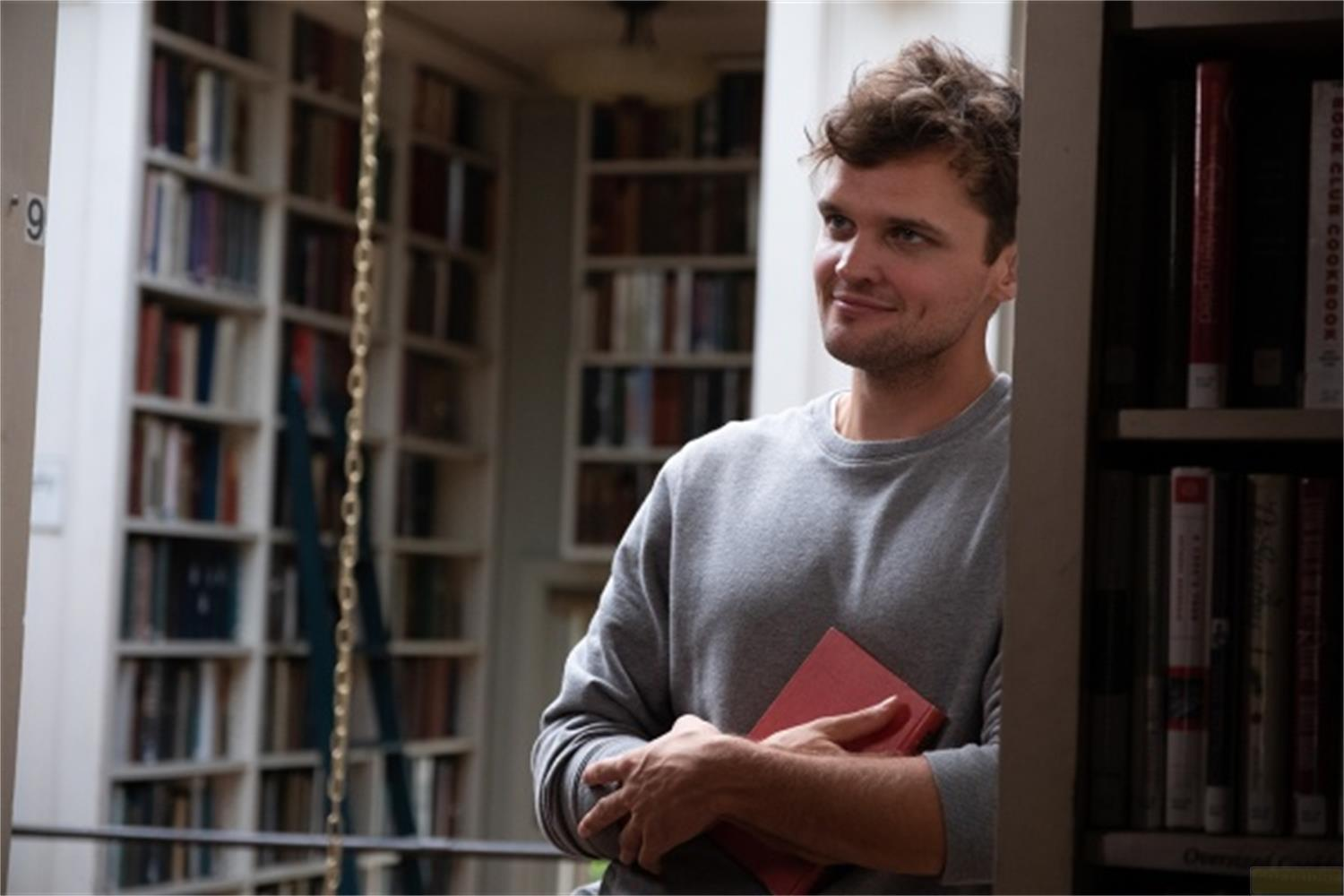 Un uomo in felpa grigia tiene un libro rosso in una biblioteca.
