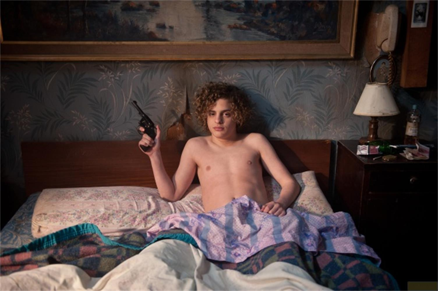 Un ragazzo a letto tiene una pistola. Sopra di lui un quadro sul muro.