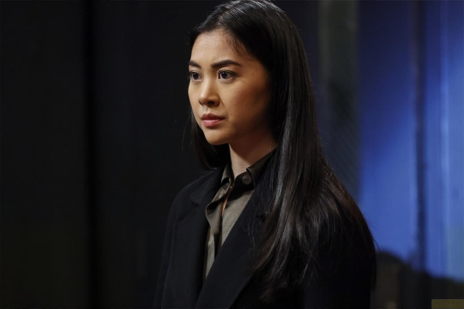 Una giovane donna con lunghi capelli neri indossa un blazer nero sopra una camicia verde oliva.