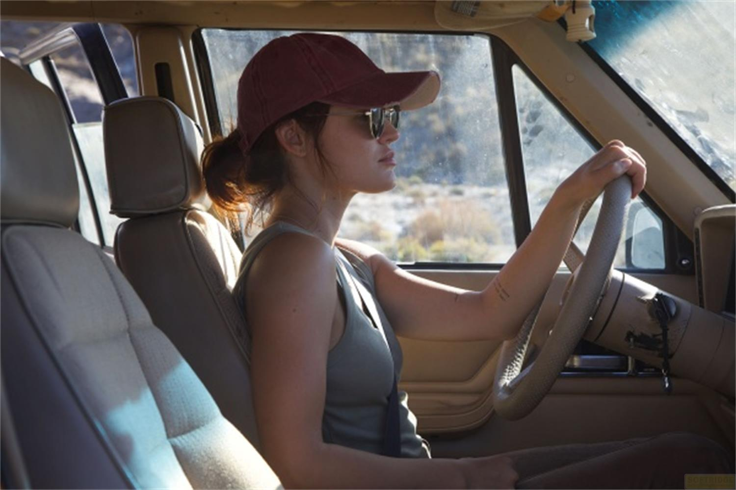 Una donna con cappellino rosso e occhiali da sole guida un'auto in un paesaggio roccioso.