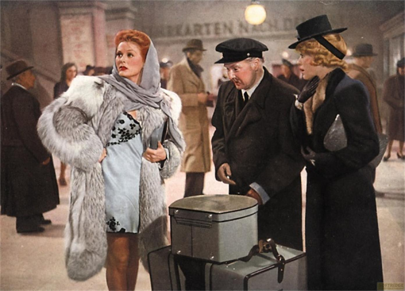 Eine Frau in Pelzmantel und Kleid steht neben Gepäck. Ein Mann in Uniform blickt sie an.