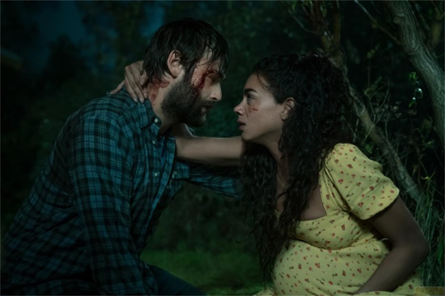 Un uomo ferito con la barba e una donna incinta si guardano nell'oscurità.