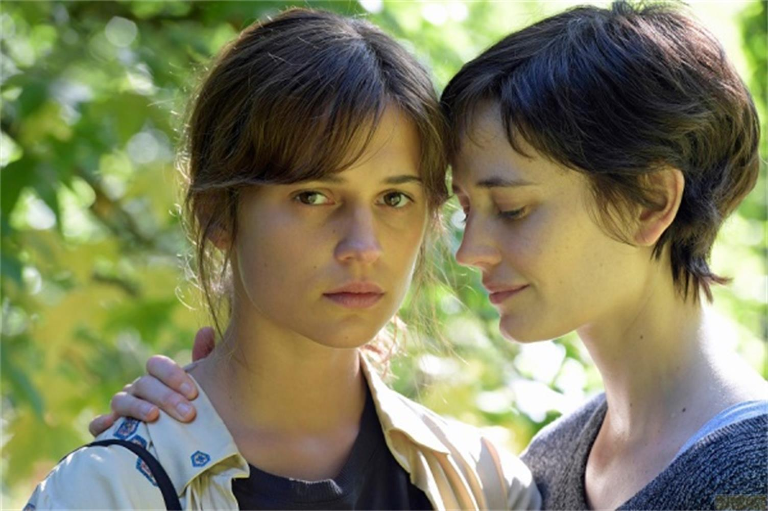 Due donne all'aperto, una che abbraccia l'altra. Foglie verdi sullo sfondo.