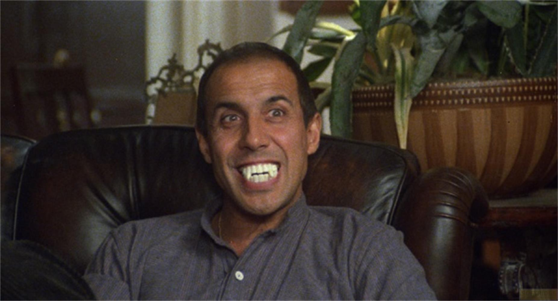 Un uomo con zanne da vampiro sorride ampiamente, seduto su un divano di pelle.