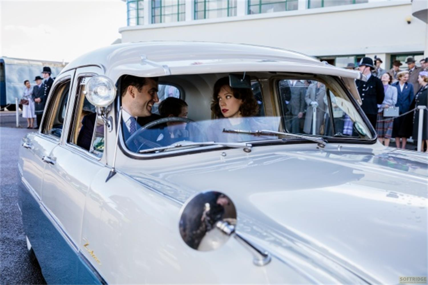 Un uomo e una donna in un'auto d'epoca; poliziotti e curiosi all'esterno.