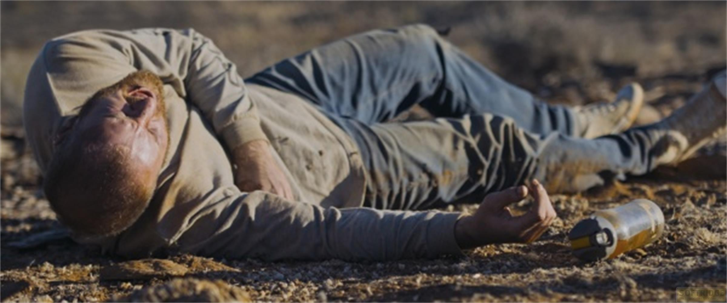 Un uomo giace privo di sensi per terra nel deserto, vicino a una bottiglia.