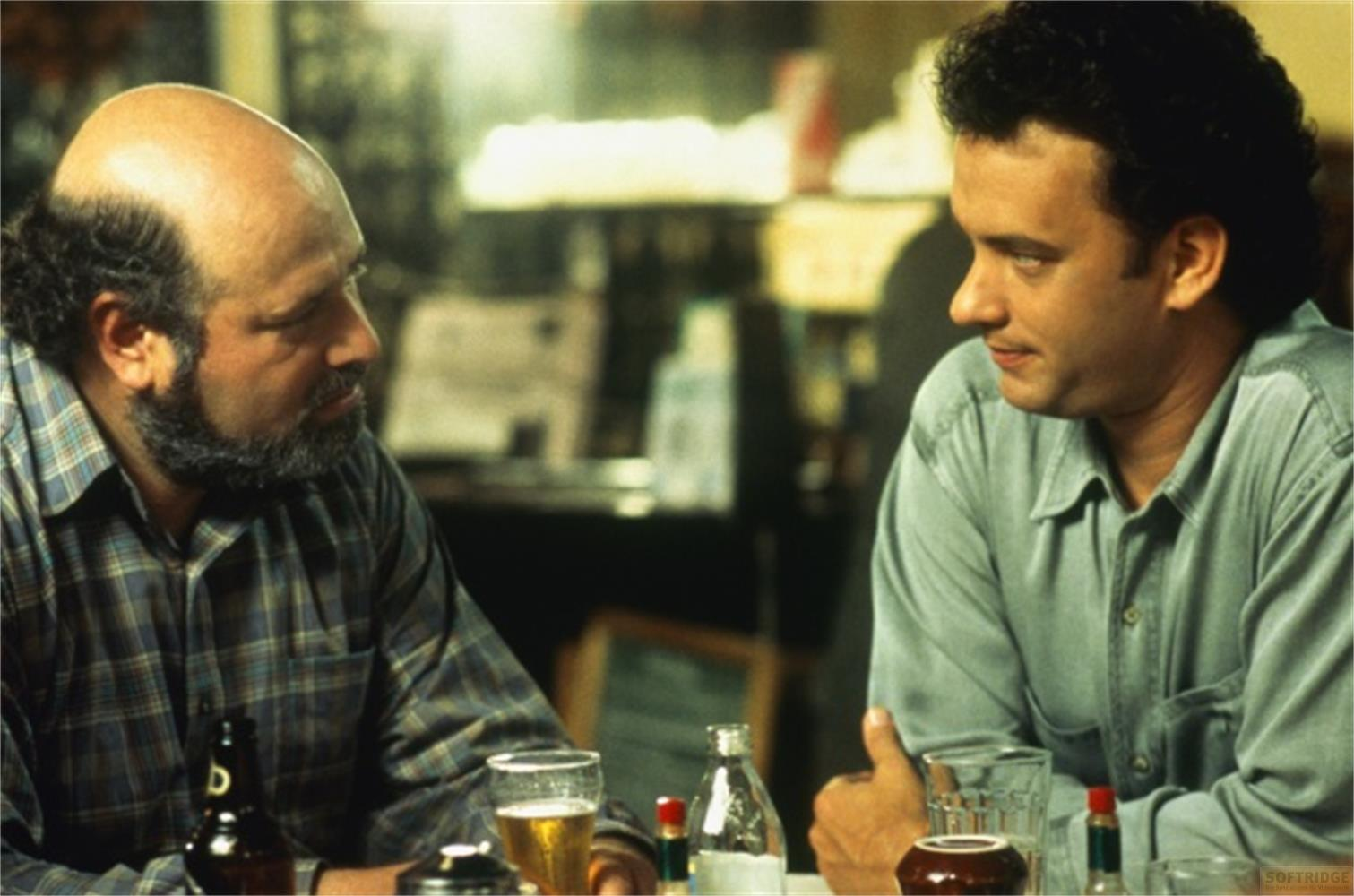 Deux hommes assis à une table. L'un est chauve avec une barbe, l'autre a les cheveux sombres.