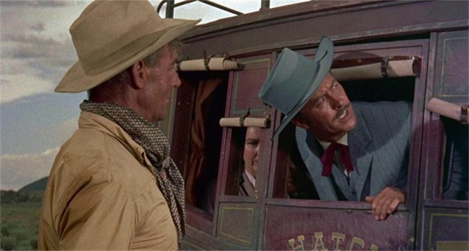Uomo con cappello parla a uomo in carrozza. Paesaggio esterno visibile.