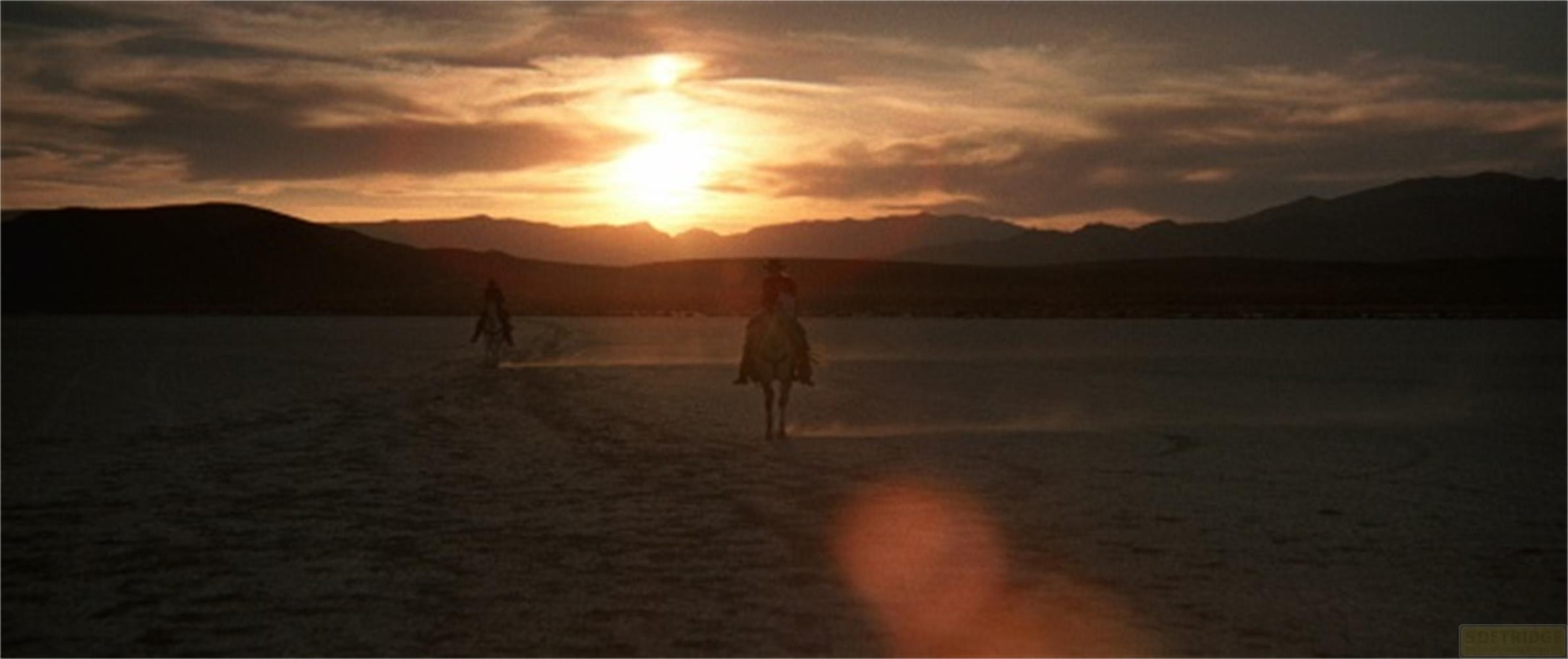 Zwei Reiter reiten durch eine staubige Ebene bei Sonnenuntergang.
