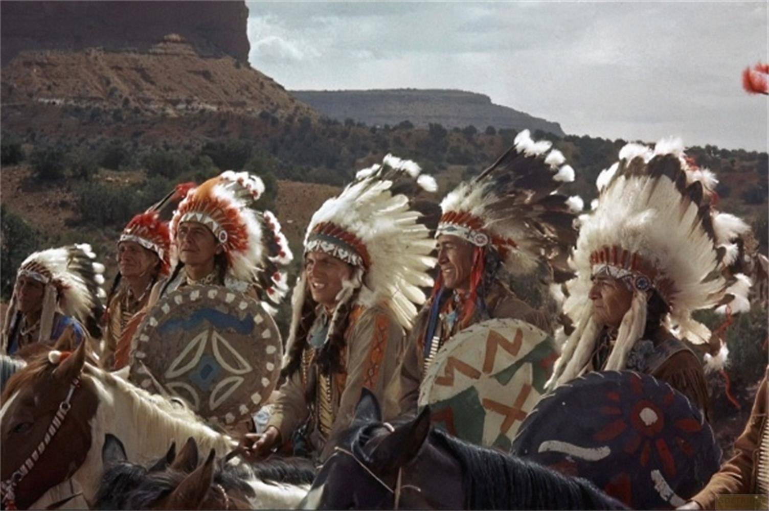 Diversi nativi americani con copricapi piumati cavalcano cavalli davanti a un paesaggio roccioso.