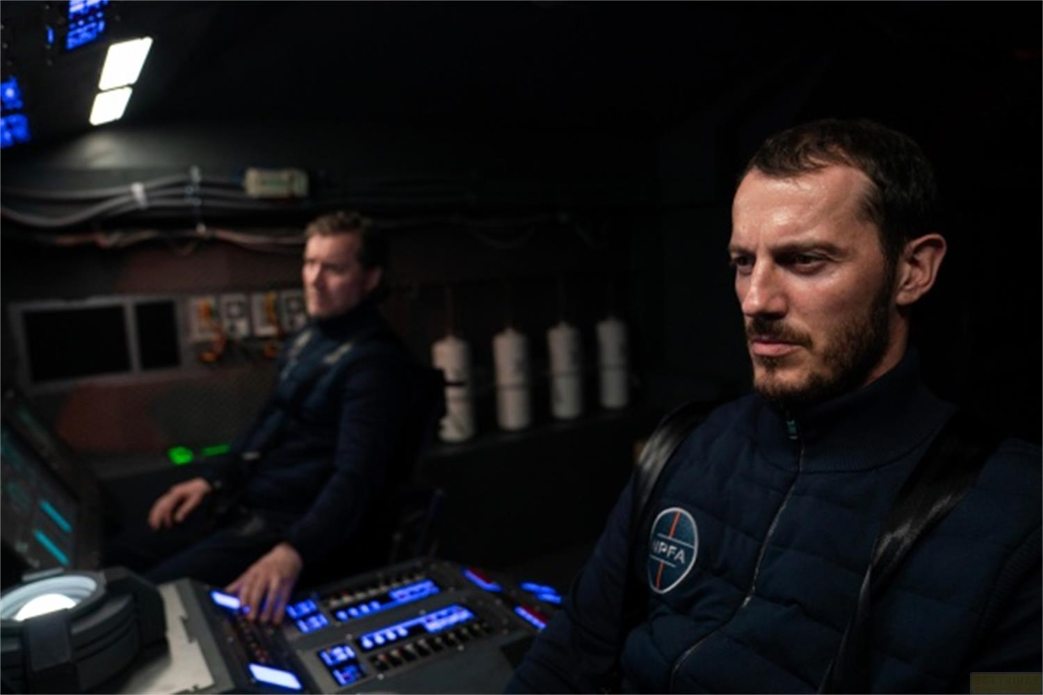 Due uomini in uniformi scure seduti nella cabina di pilotaggio di un'astronave con pulsanti.